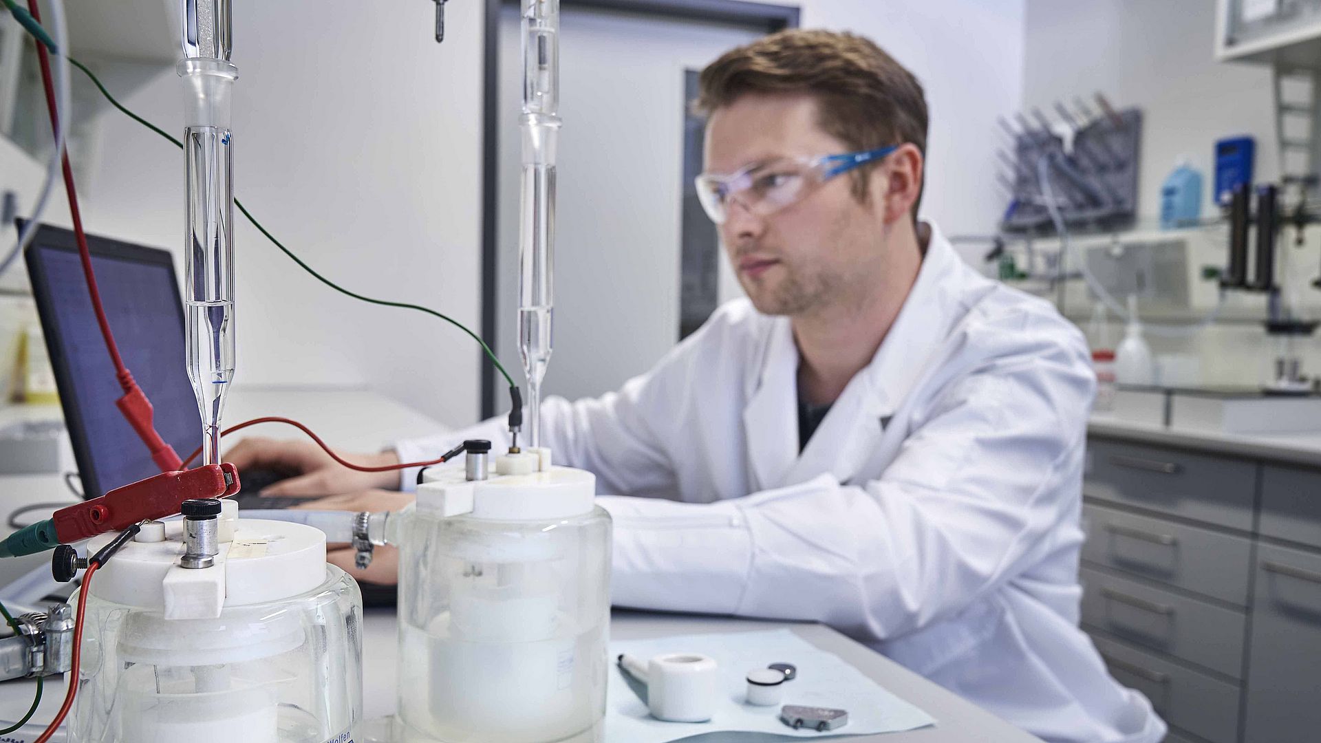 A man with safety goggles and a lab coat sits in a laboratory